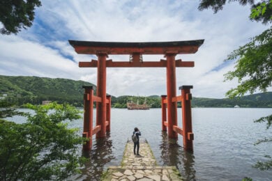 A person stands on a stone path under a large red torii gate by a lake, with green hills and a ship in the background under a partly cloudy sky.